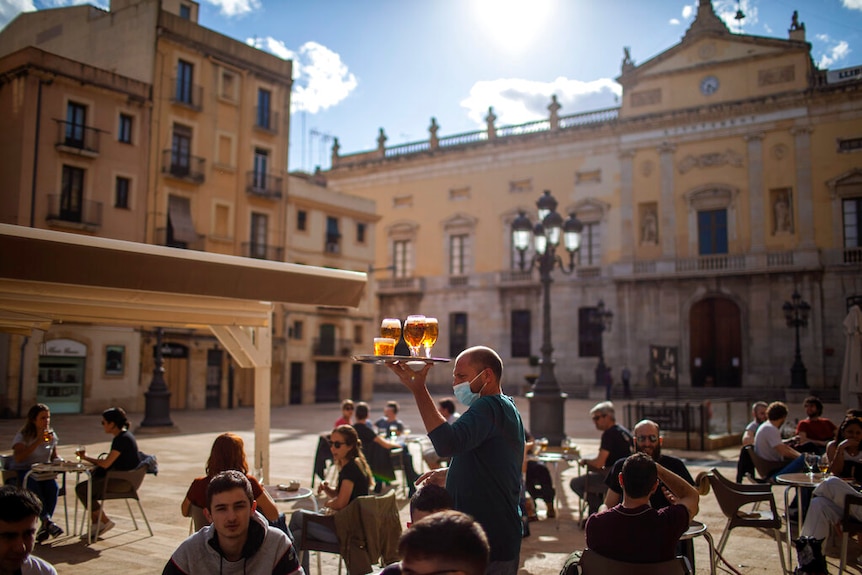 You view people sitting in an open plaza as a waiter carries beers in wide stemmed glasses on a silver plate.