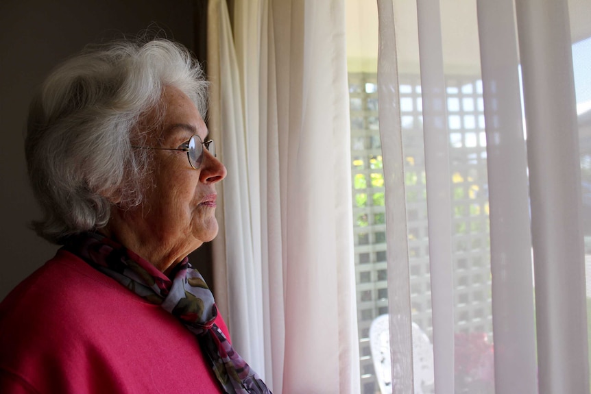 An elderly woman stands at a window and looks out.
