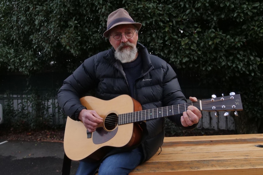по-възрастен мъж със сива брада и шапка, свирещ на китара 