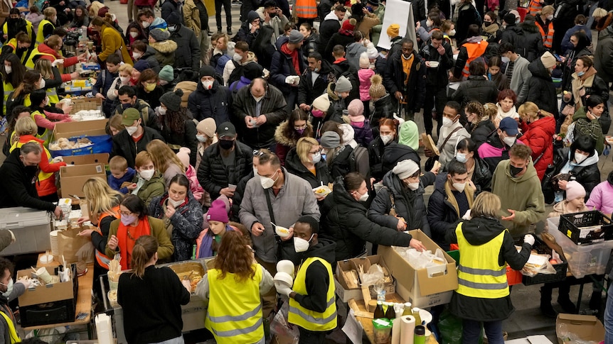 A huge crowd of refugees waits for food to be distributed from volunteers who stand behind tables in a large train station.