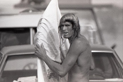a black and white photo of a man dripping wet putting his surfboard back into its cover standing next to a car