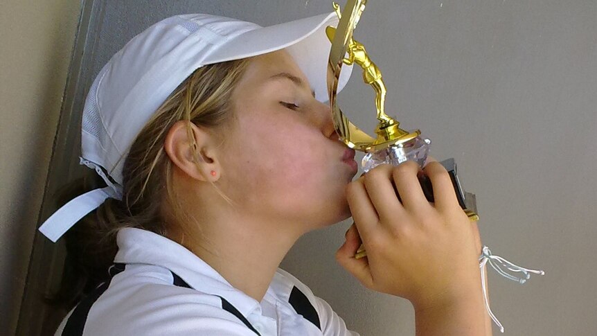 A girl wearing a polo shirt and cap holds a trophy, kissing it.