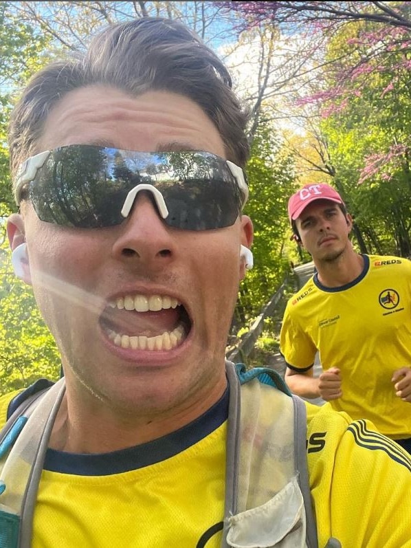 A man taking a selfie while running. He is wearing dark sunglasses, a yellow Australia top and is followed by another runner.