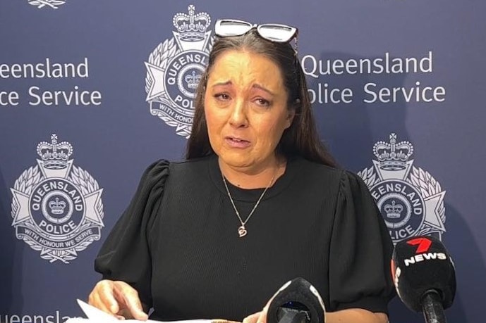 A woman in a dark dress, looking upset.  She is standing in front of a dark blue Queensland Police Service banner.