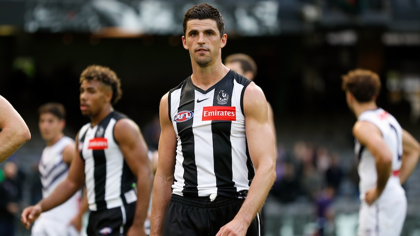 A Collingwood AFL player walks while teamates and opposition players are behind him.