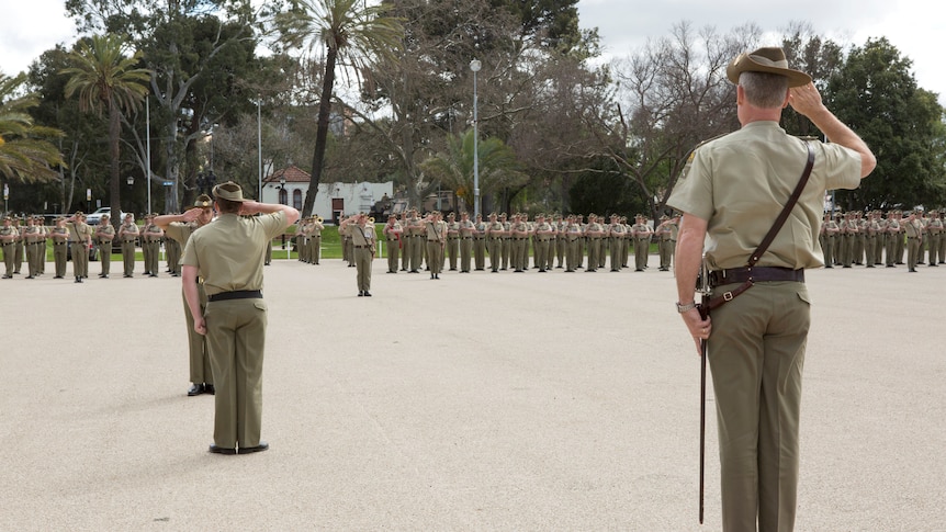 An army leader saluting in khaki on a platform in the foreground, with a long row of younger soldiers in the background