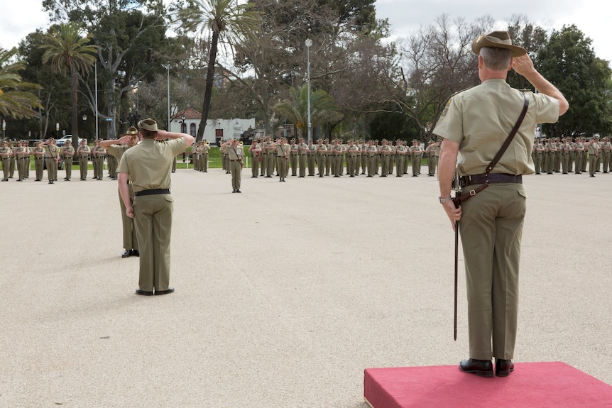 An army leader saluting in khaki on a platform in the foreground, with a long row of younger soldiers in the background