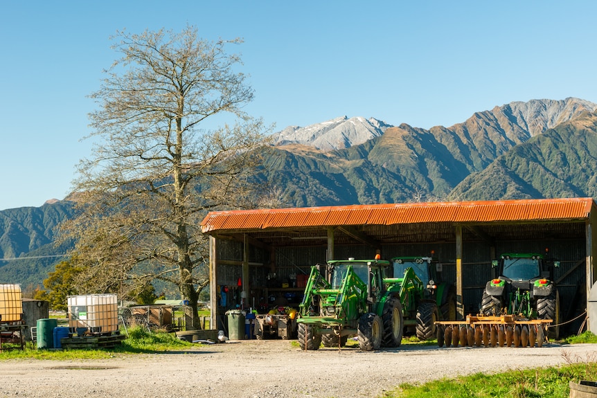 A farming shed with tractors is seen with the alps in the background. 