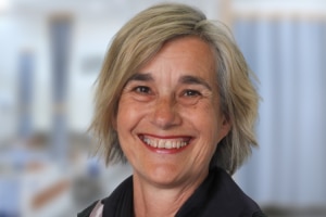 A middle aged white woman with short chin length grey hair and wearing a black top, smiling in front of a blurred background.