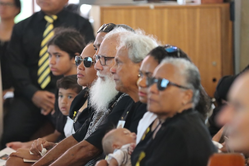 A row of people sitting on a church pew, dressed in black and looking serious.
