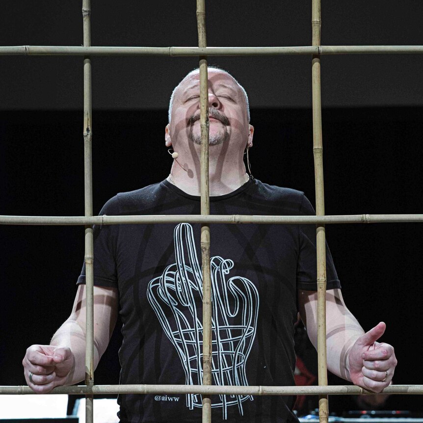 Nikolai Khalezin stands on stage behind wooden bars, his eyes closed and his head raised.