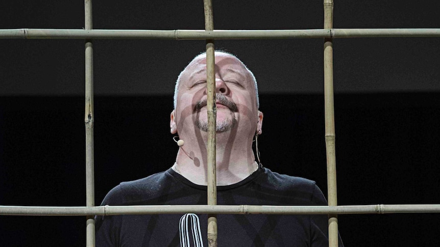 Nikolai Khalezin stands on stage behind wooden bars, his eyes closed and his head raised.