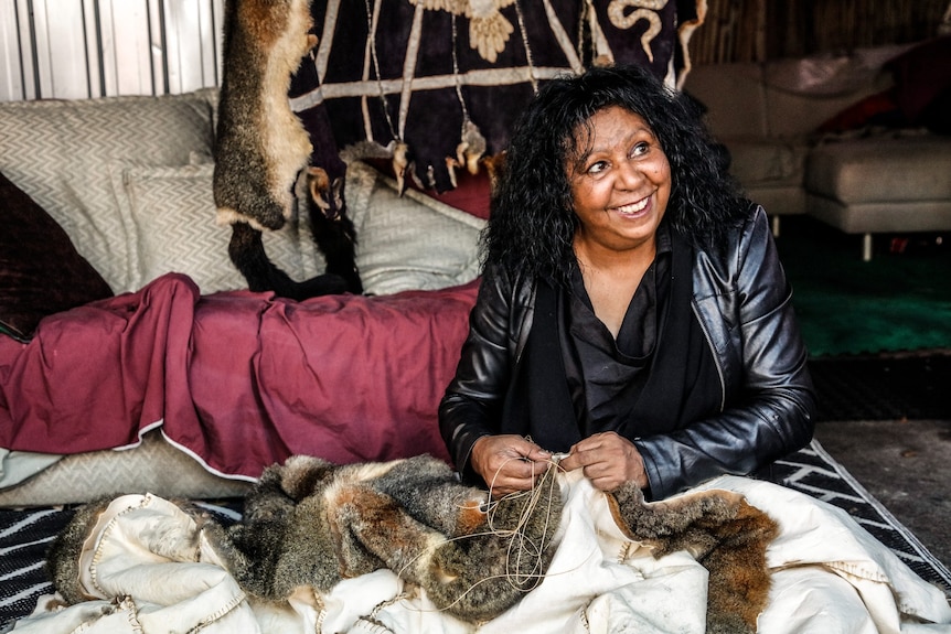A 60-something Aboriginal woman with a big smile on her face sews a possum skin cloak