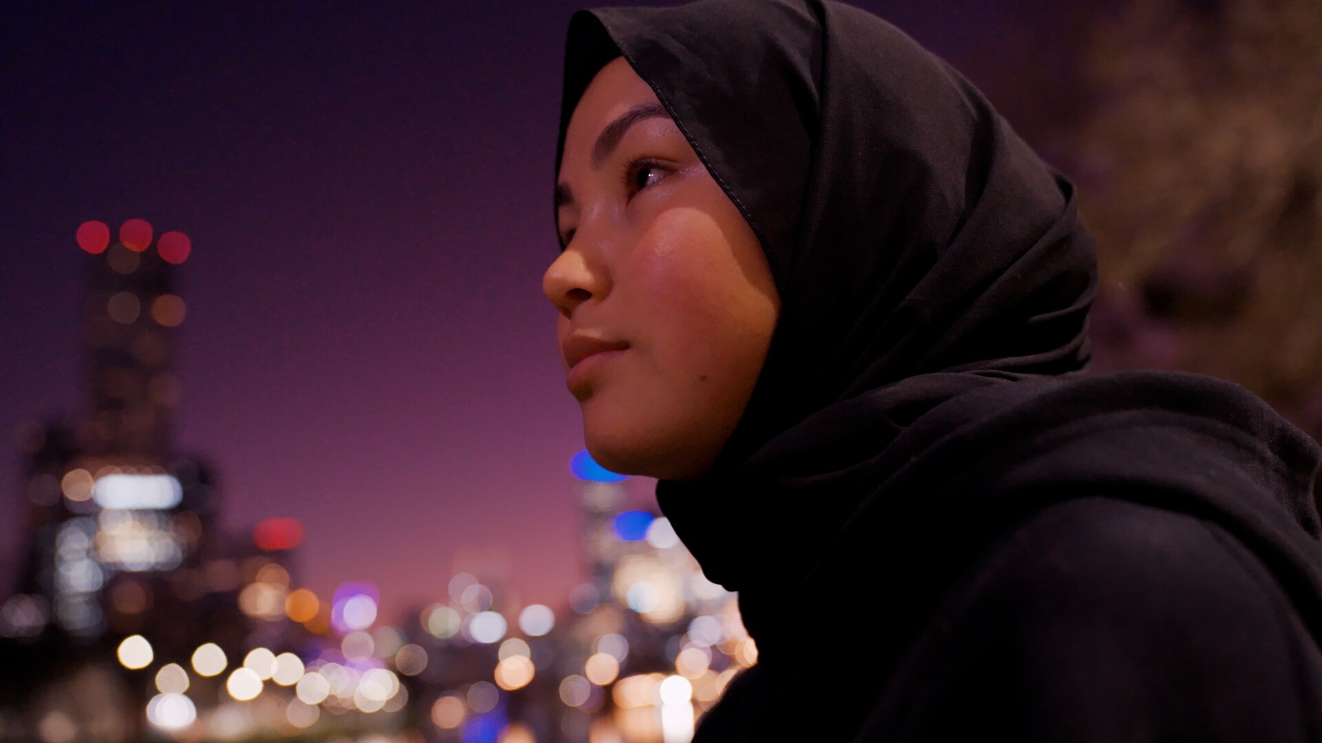 Adiba looks out across the Melbourne city in the evening with blurred city lights in the background