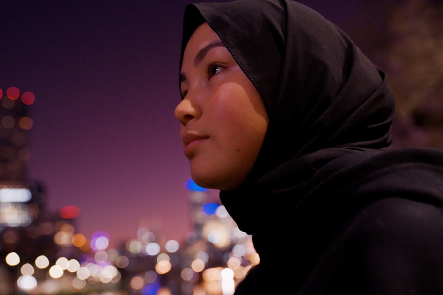 Adiba looks out across the Melbourne city in the evening with blurred city lights in the background