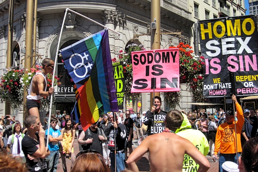 Pro and anti gay activists clash during protests on a street corner, with one placard that reads 'Sodomy is Sin'