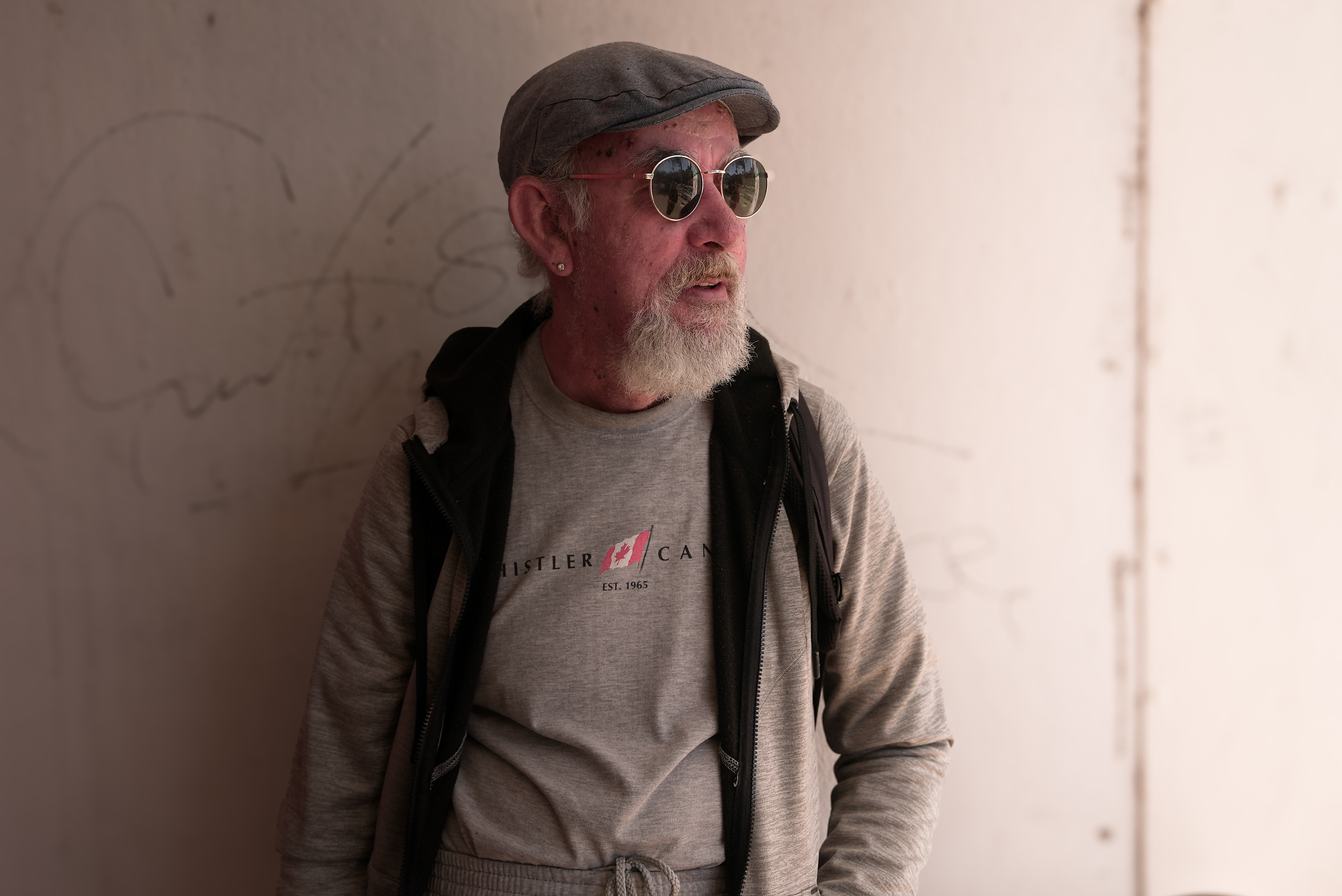 A man with a grandpa hat on and sunglasses leans against the wall and looks off to the right