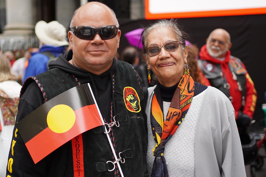 A man and woman stand close together with an Aboriginal flag in front of them
