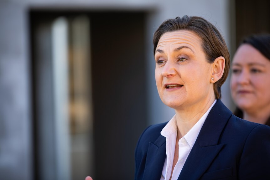 A woman with short hair and a blue blazer speaks.