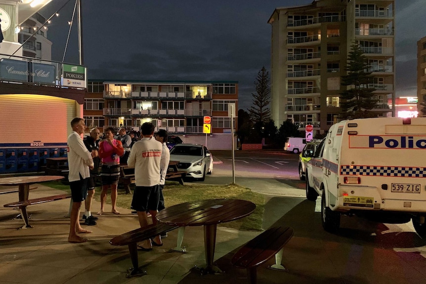 Middle-aged men in shorts and fold their arms and engage in discussion at night near lit buildings,