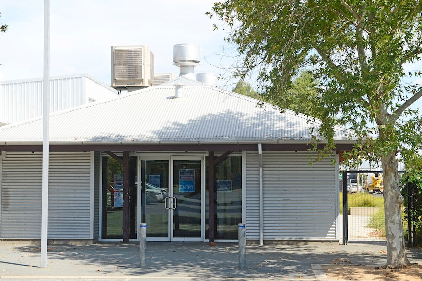 A light-coloured, single-storey building shaded by a tree on a sunny day.