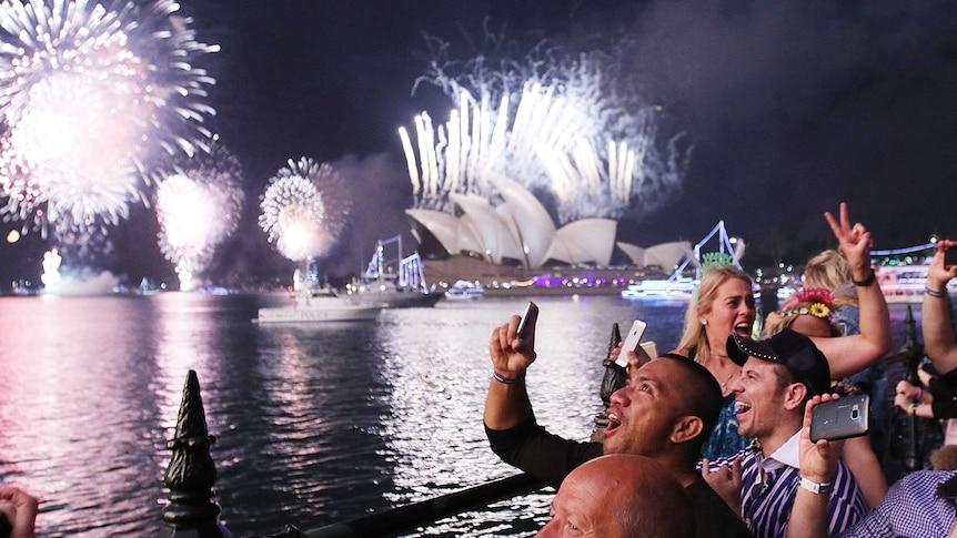 A group of people celebrate next to the water, with Sydney Opera House in the background lit up by fireworks.