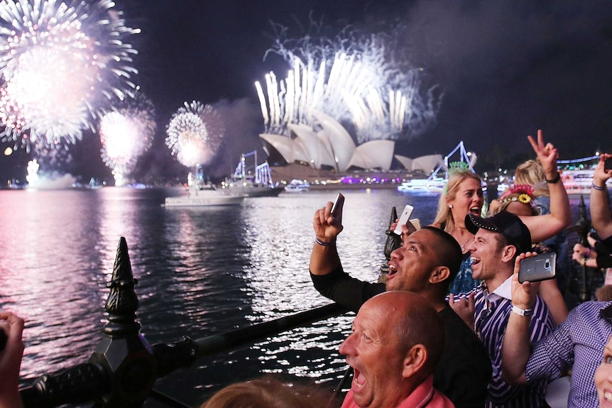 A group of people celebrate next to the water, with Sydney Opera House in the background lit up by fireworks.