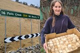Restored landsip site with a street sign saying Falls Creek/Mount Beauty and a young woman holding a box of dirt cubes.