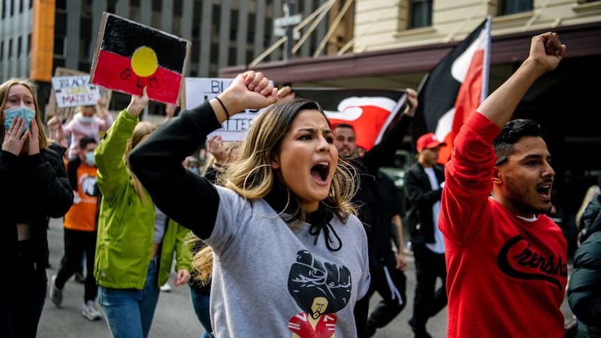 A woman and a man raise their fists towards the air, shouting, as part of a crowd waving placards and Aboriginal flags