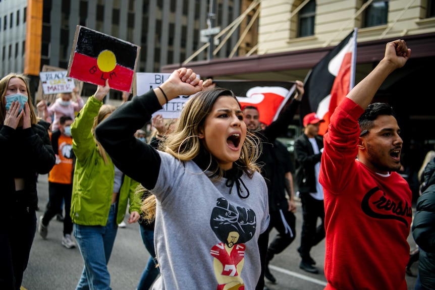 A woman and a man raise their fists towards the air, shouting, as part of a crowd waving placards and Aboriginal flags