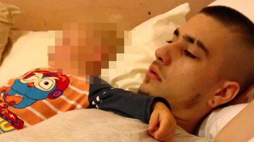 A young man in a grey t-shirt lies on a bed with a baby wearing an orange striped top, whose face is pixelated.