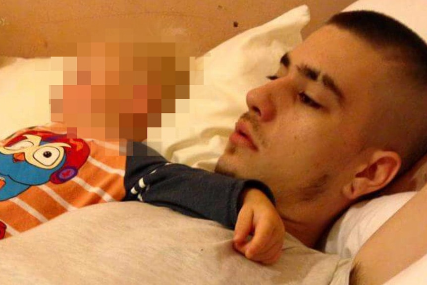 A young man in a grey t-shirt lies on a bed with a baby wearing an orange striped top, whose face is pixelated.
