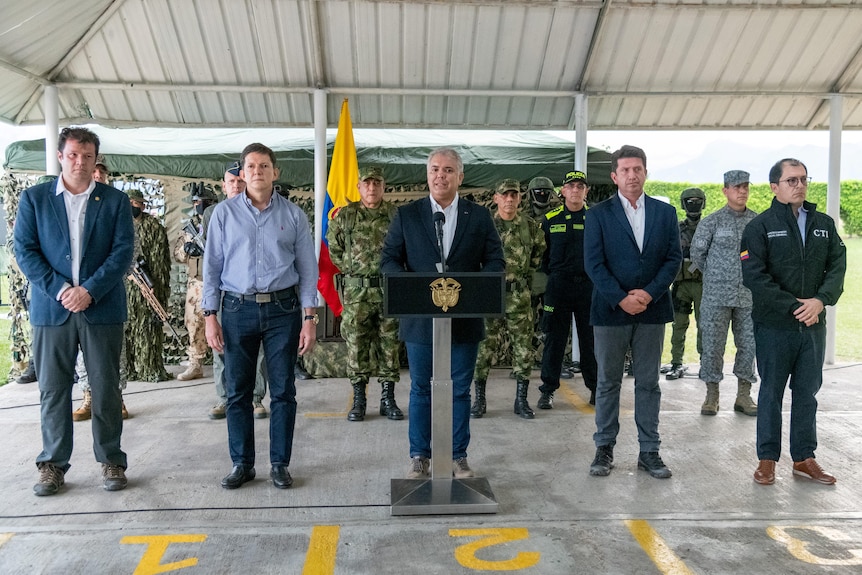 A man stands at a podium addressing the camera, with other officials and army surrounding him.