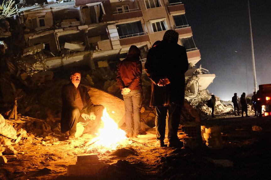 People huddle round a fire, surrounded by ruins.