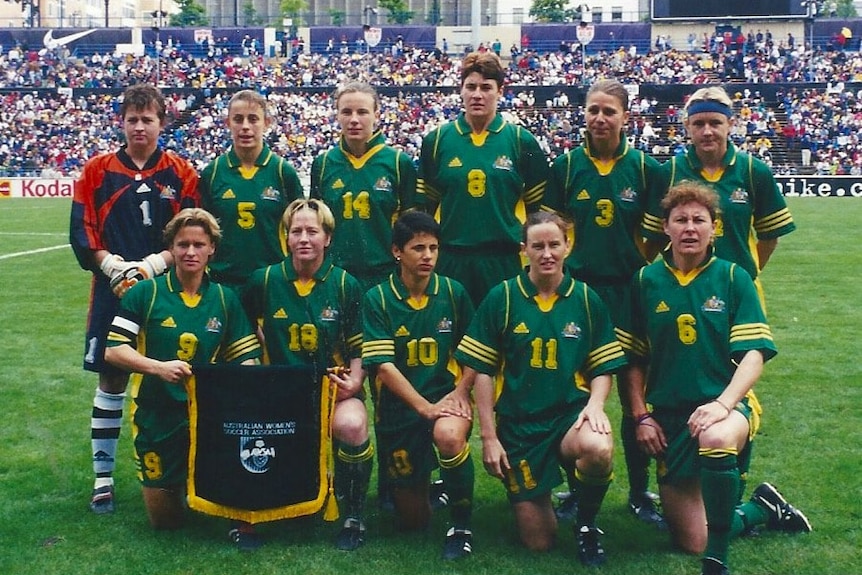 A women's soccer team wearing green and yellow poses for a photo before a game with a crowd in the background