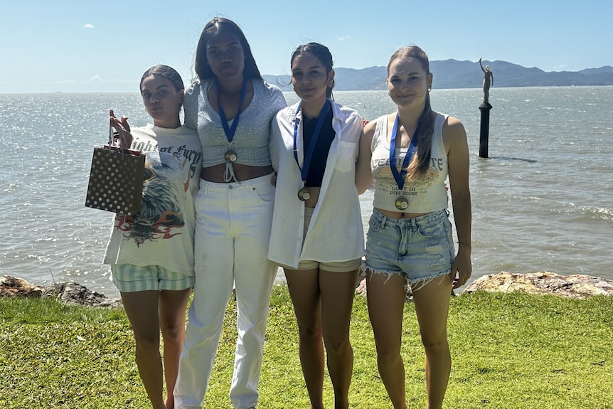 Four young women pose with their arms around each other, smiling. Three of them are wearing medals.