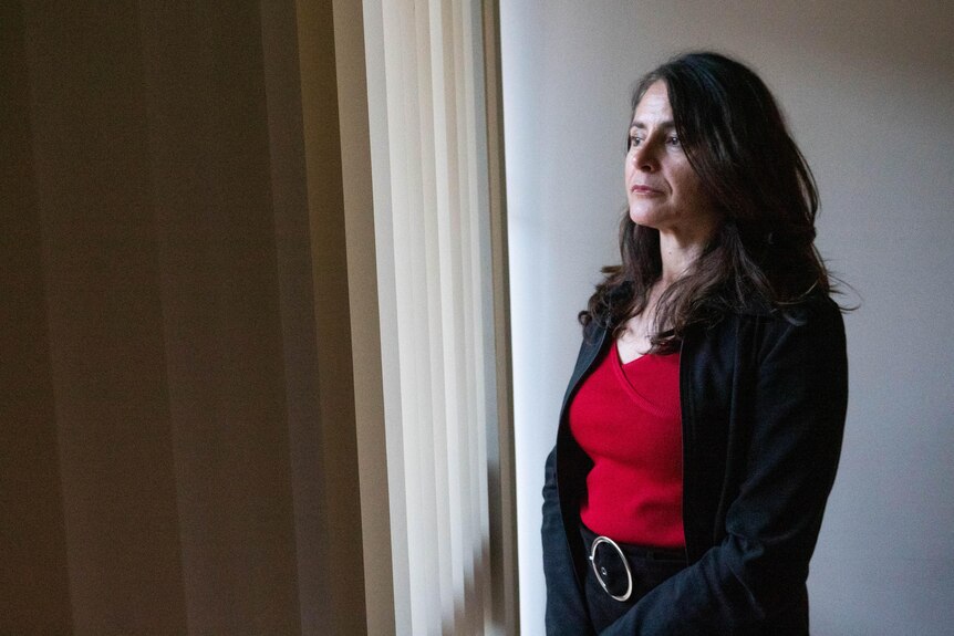 Nicki Petrou looks out a window. The dark-haired woman is wearing a red top and a black cardigan