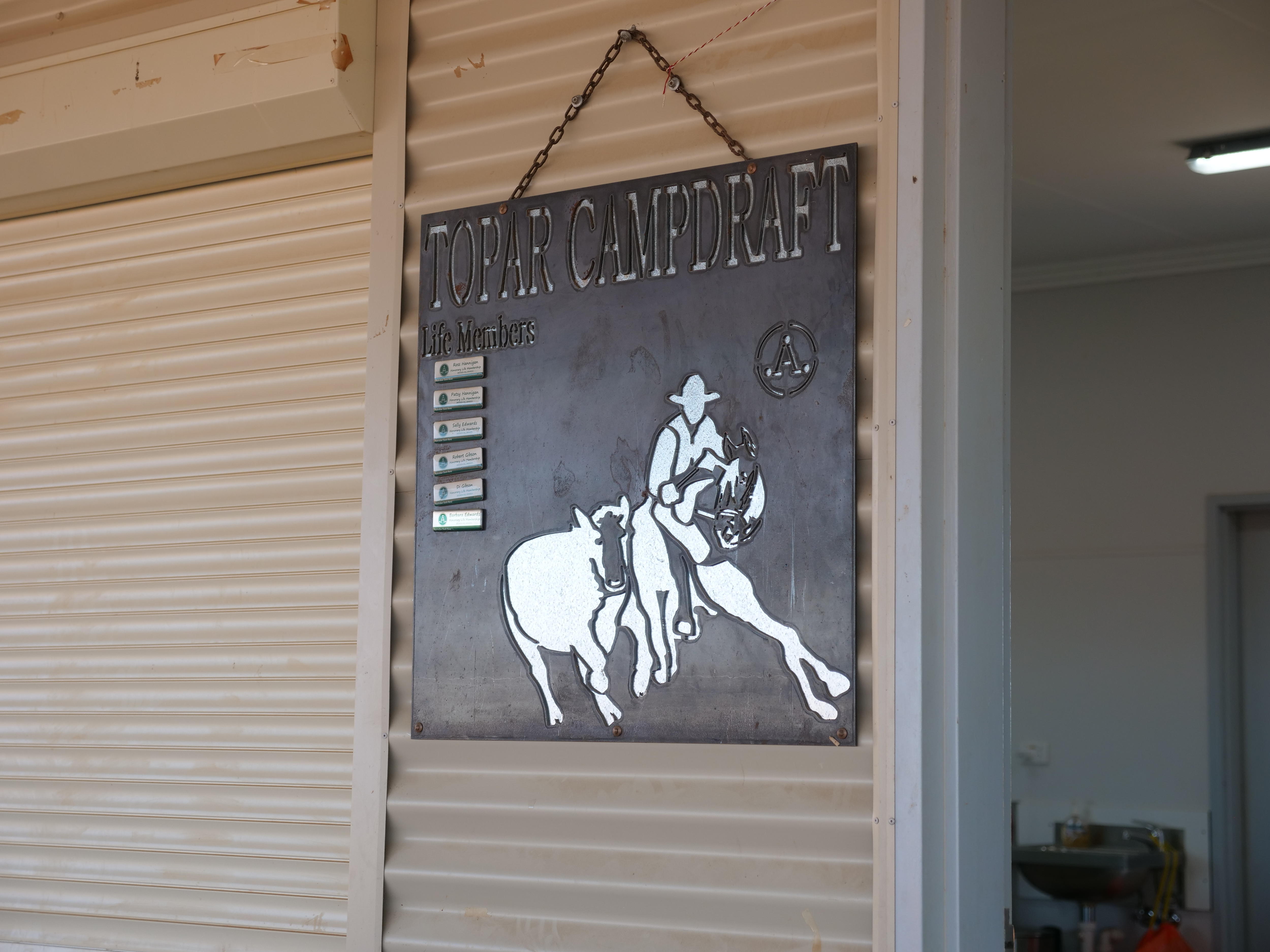 black sign with a horse and cow cut out of it on a white tin wall that reads Topar campdraft 