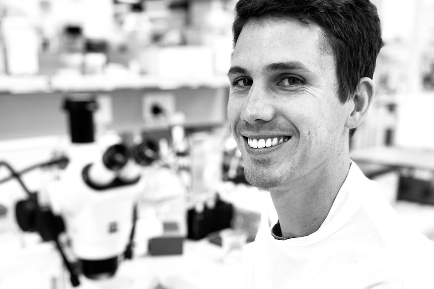 Black and white photo of a young, dark-haired man sitting beside a microscope smiling