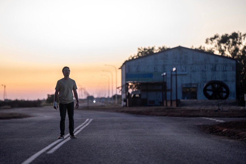 A man walks on a dusty outback road in front of a shed and a sunset.