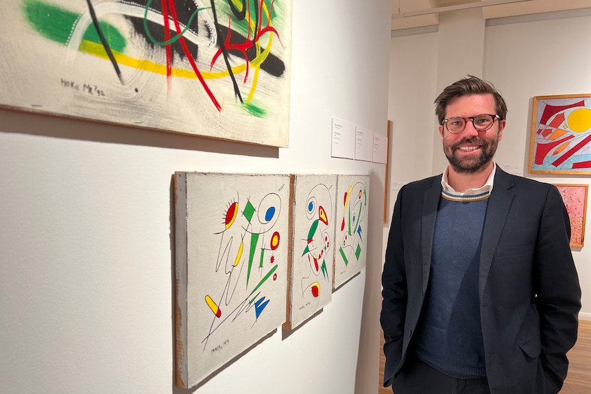 A middle-aged healthy man with brown hair and beard stands next to modernist paintings in gallery, smiling