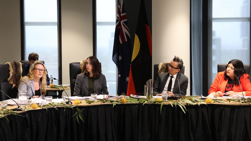 Four people sit at a table adorned with gum leaves, with flags behind them.