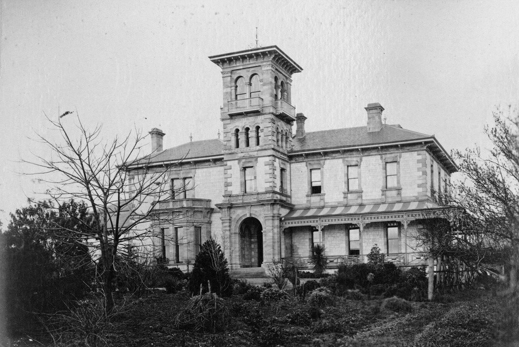 Uma fotografia em preto e branco de uma mansão, com arbustos e árvores mortas na frente.