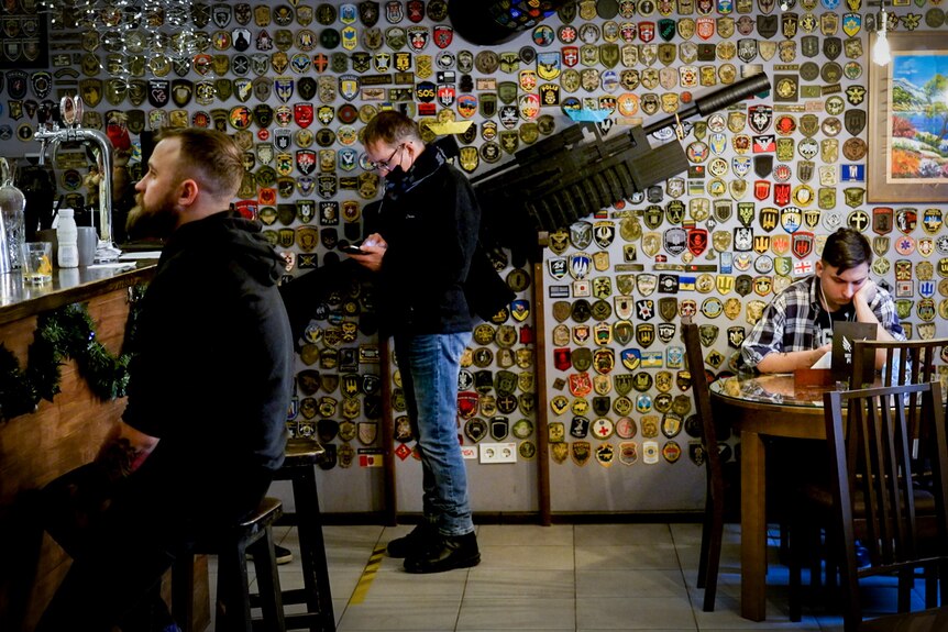 Three men sitting in a pizza shop with a massive gun hung on the wall behind them