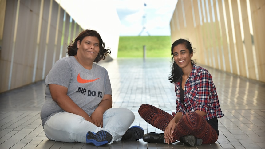 A young man and young woman sit on the ground in the foreground of Parliament house, Canberra.