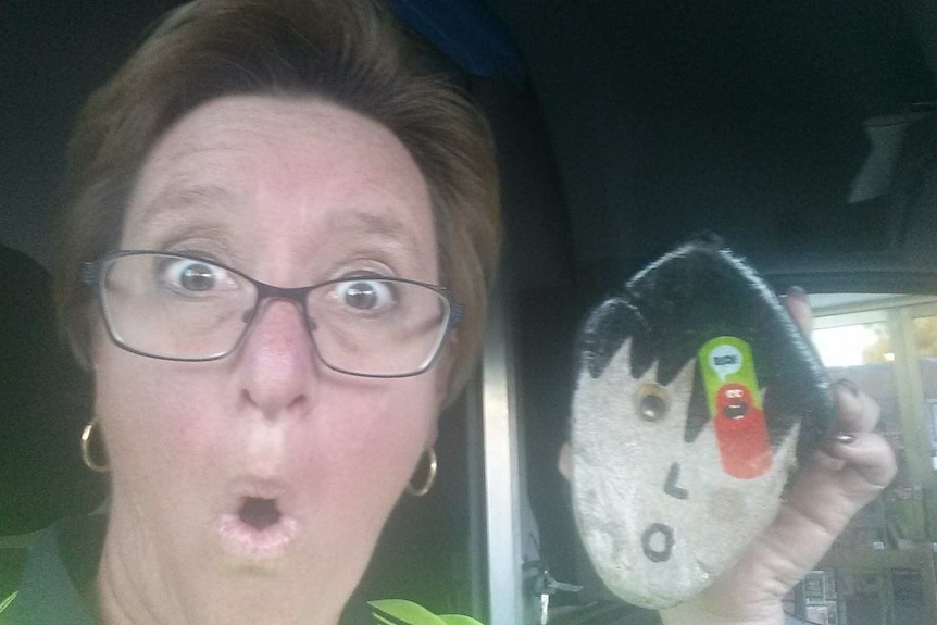 A woman with short brown hair and glasses holding a pet rock next to her face.