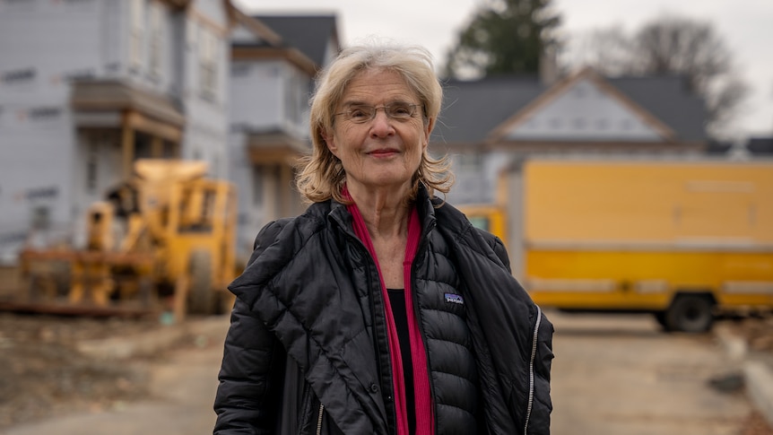 An older woman wearing a black puffer jacket and maroon jumper stands in front of a construction site