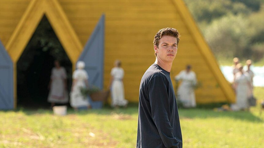 Will Poulter is standing in a green field with a yellow structure behind him