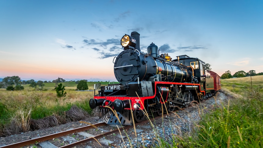 A black steam engine with red trim pulls a red carriage through the grassy countryside at dusk. 