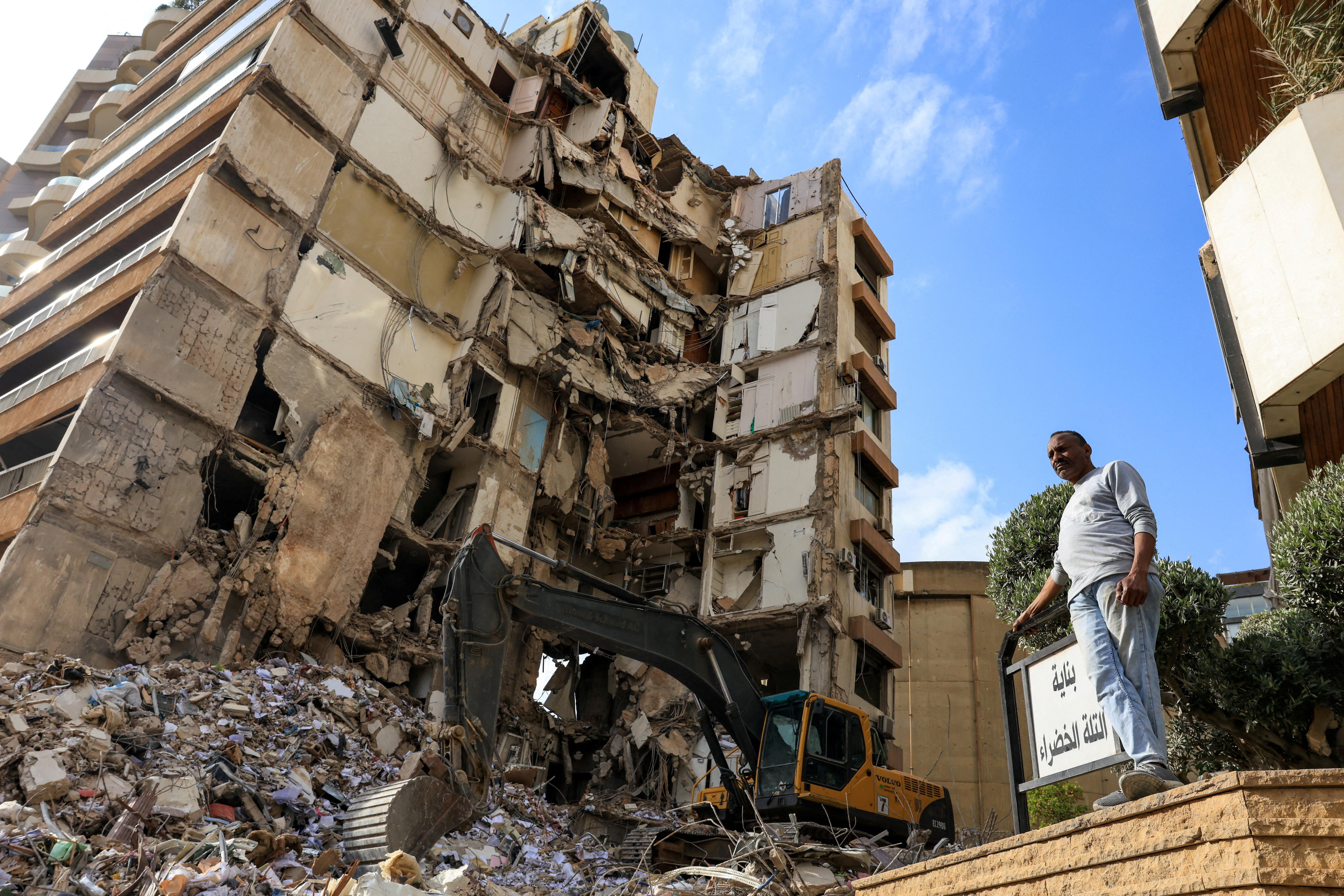 A man stands near a damaged building from which piles of rubble spill to the ground